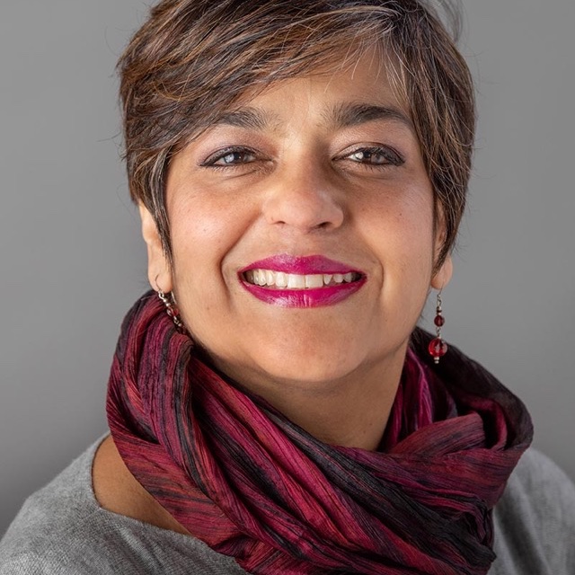 A person with short brown hair and pink lipstick wearing a gray shirt and black and red scarf against a gray background, smiling at the viewer.