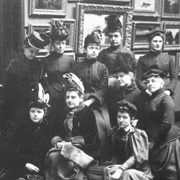 A monochrome historical photo of a group of people in old-fashioned dresses and formal hats, looking at the camera.