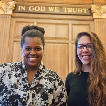 Two people stand together in front of an ornate wooden door with "In God We Trust" above it. The person on the left has black curly hair in a bun and a floral shirt. The person on the right has long straight brown hair, glasses, and a black shirt.