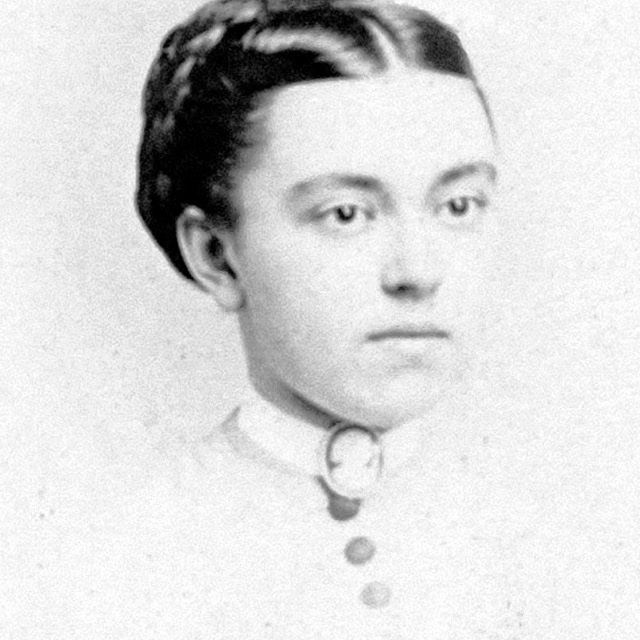 A monochrome historical photo of a person with an old-fashioned button-down dress and black curly hair arranged into a bun.