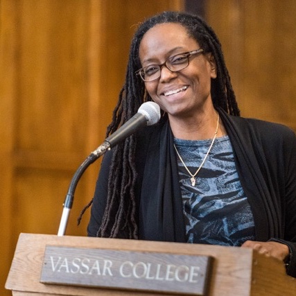 A person with long black braided hair and glasses stands at a podium, smiling and speaking into a microphone.