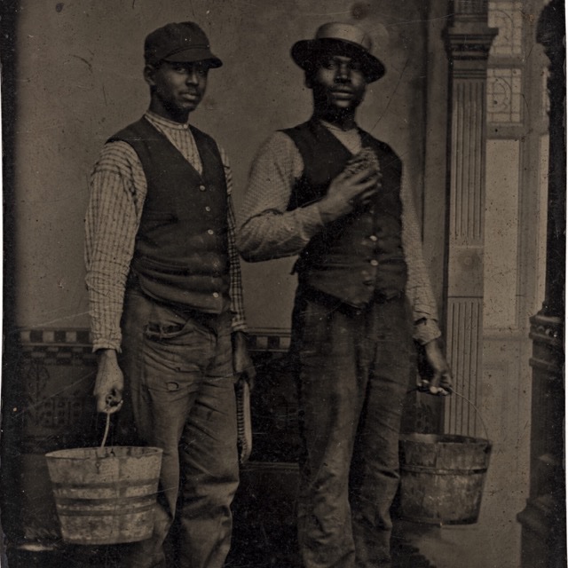 A monochrome historical photo of two people in old-fashioned clothing carrying wooden buckets.