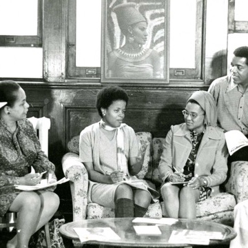 A monochrome historical photo of several people in a room sitting around a table talking.