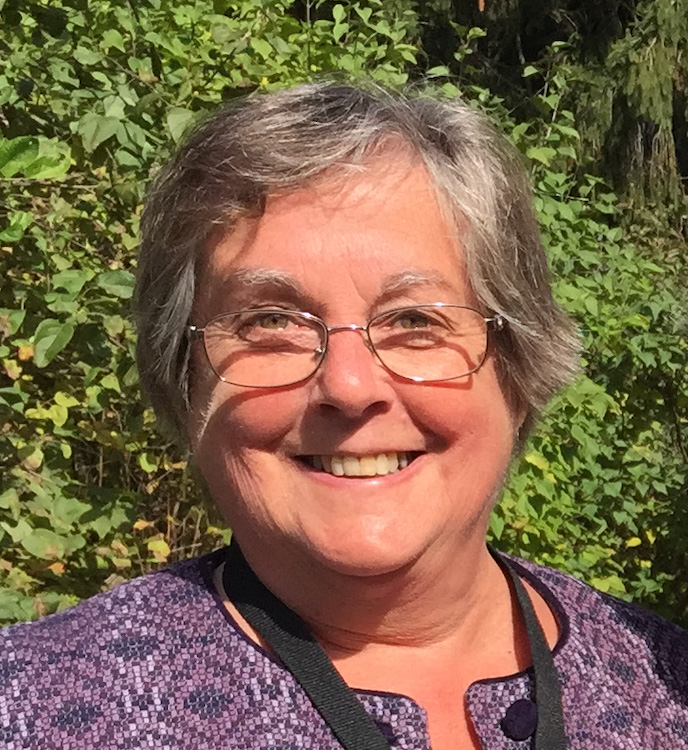 Person with glasses, purple shirt, and grey hair smiling in front of green shrubs outside.