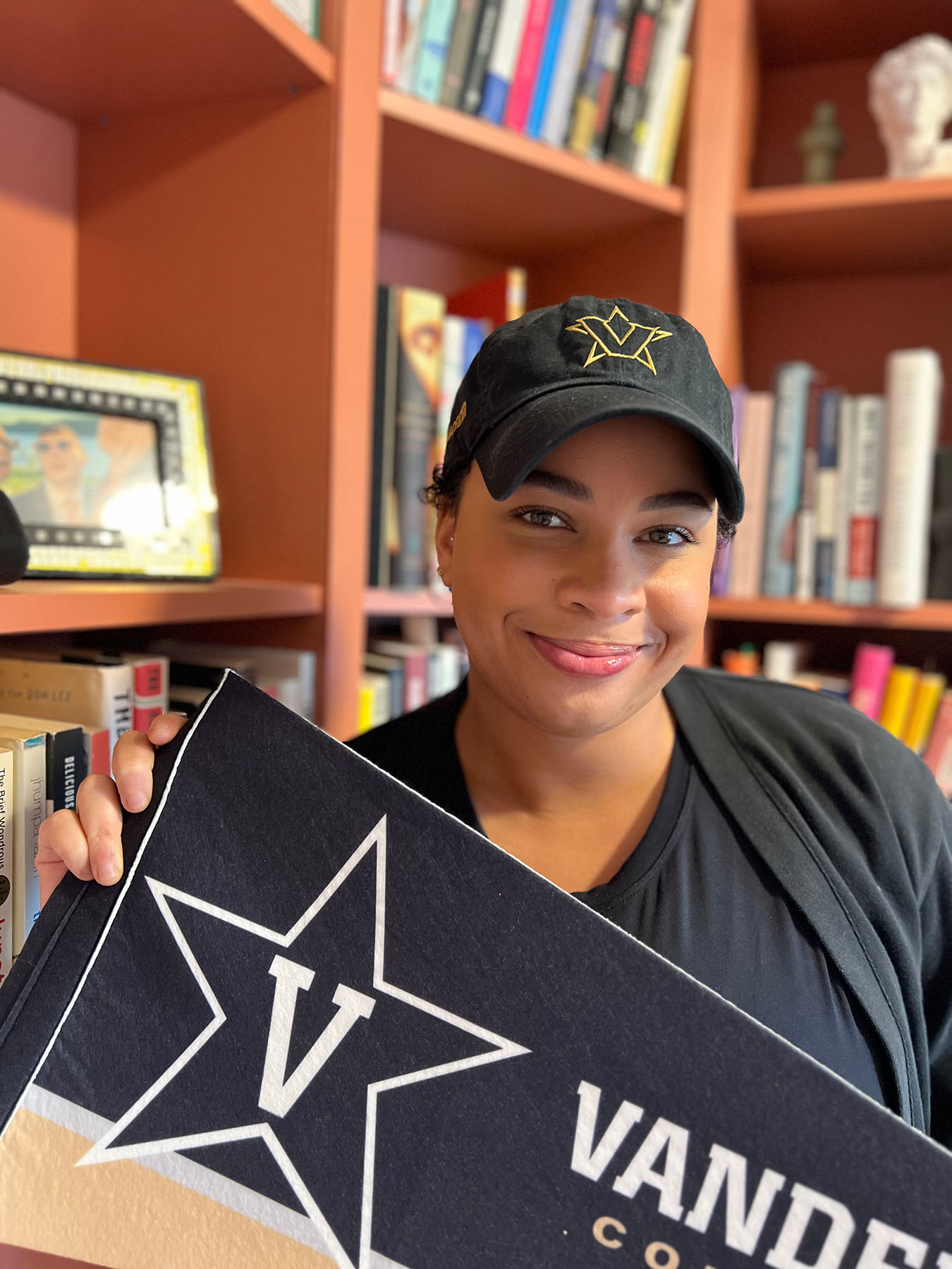 Person wearing a hat and standing in front of bookcases holding a banner/flag with a "V" pictured on it.