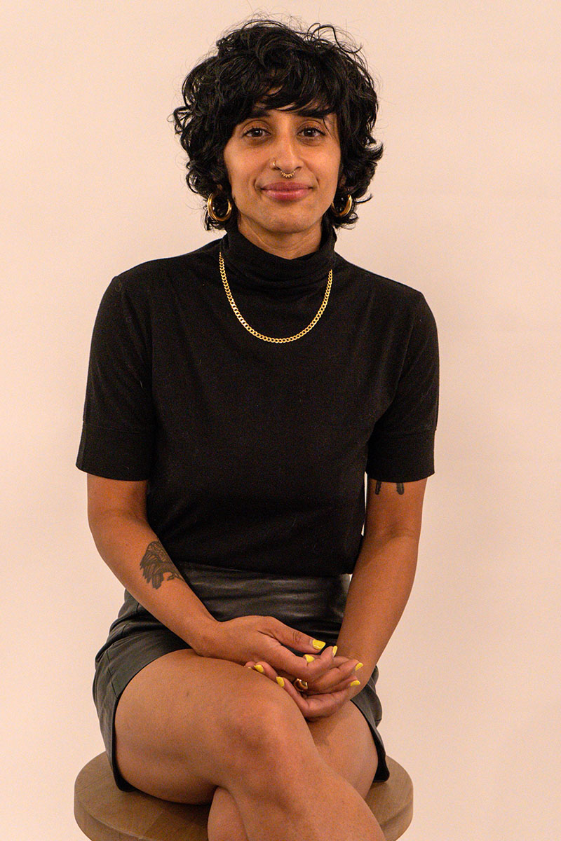 Sukaina Hirji wearing a black shirt, black skirt, and gold chain, sitting on a stool.