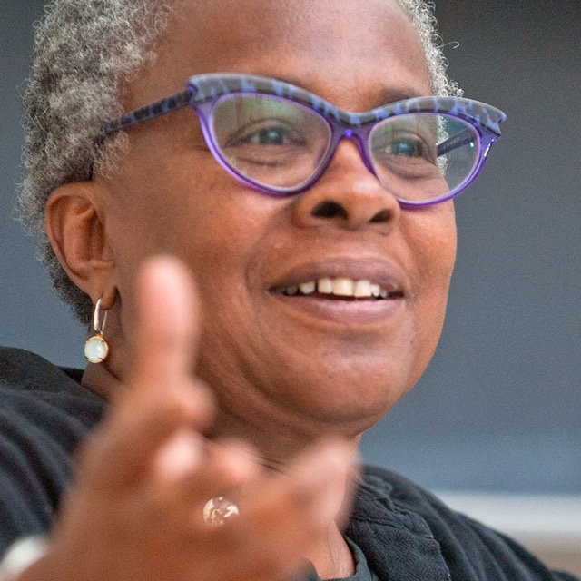 A person wearing a black shirt and purple eyeglasses with arm extended against a dark background.