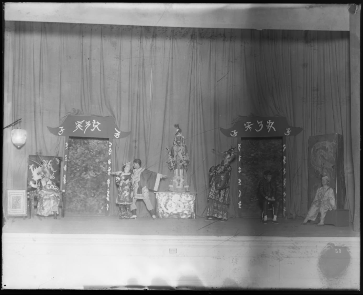 A black-and-white historical photo of a theatrical production featuring costumed actors and stage settings that are intended to look Chinese.