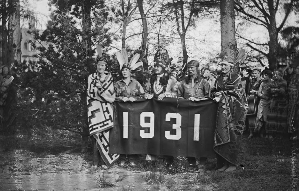 A black-and-white historical photo of Vassar students in native American outfits, standing outside and holding a large banner that says "1931" on it.