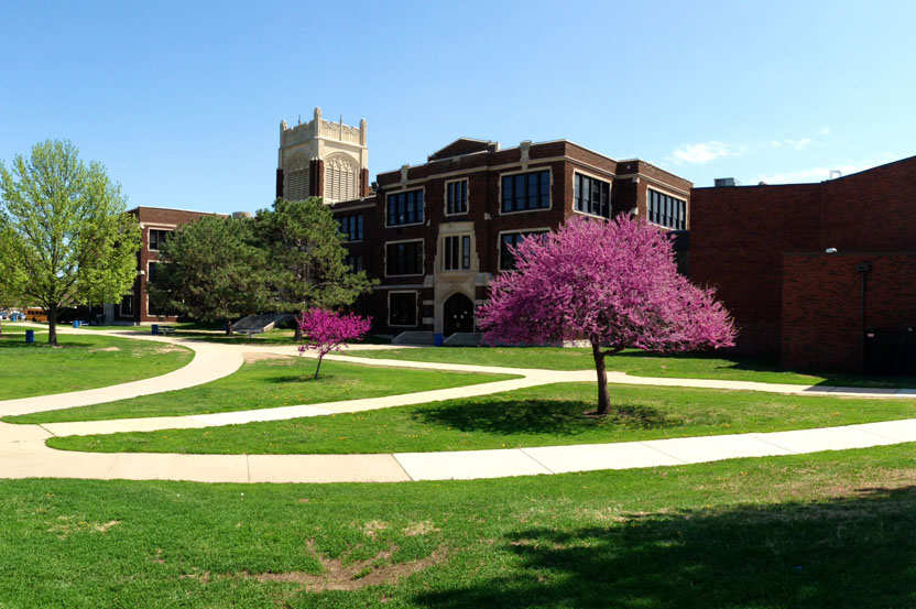 A large red-brick three-story building with a church-like tower and many windows. The building is surrounded by green grass quads.