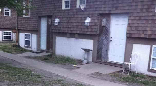 The exterior of an apartment building with white concrete walls and brown wooden shingles on the side. Doors and windows are visible. The building is surrounded by sparse grass.