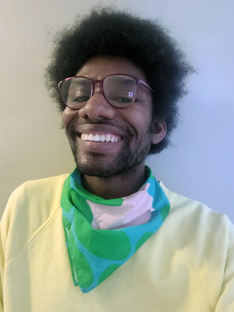 Headshot of a person with a vibrant smile wearing glasses, a yellow sweater and a scarf in teal, green, and white around the neck.