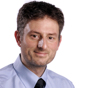 A cropped headshot of a person with dark short hair wearing a collared shirt with tie.