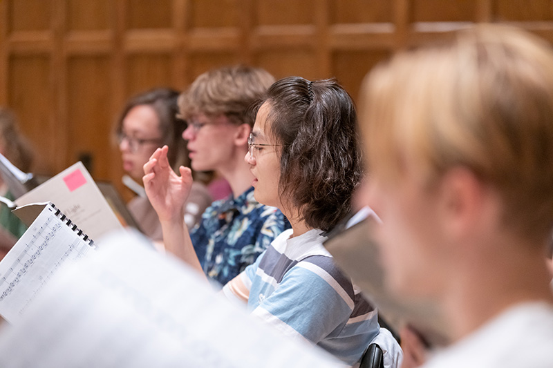 A row of singers in rehearsal showing four people, focused in the center on a person with glasses singing with her arm raised.