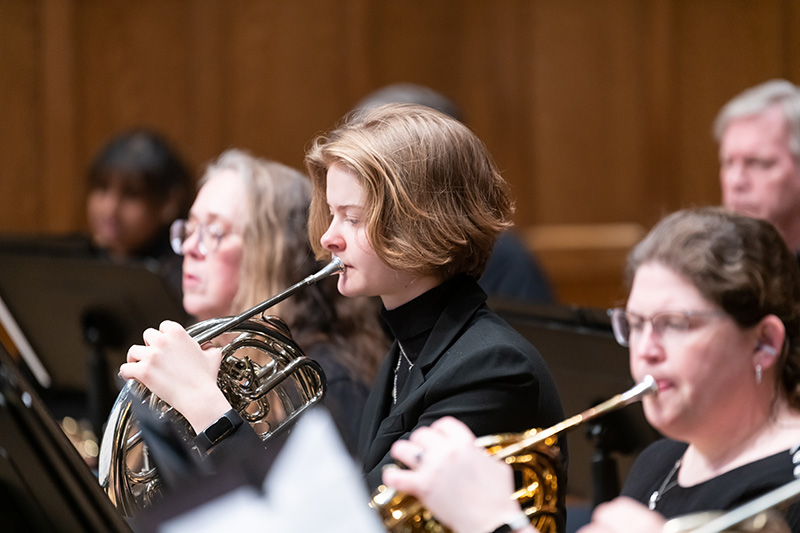 A performance, image focused on the center subject, a person with short straight hair playing the French horm.