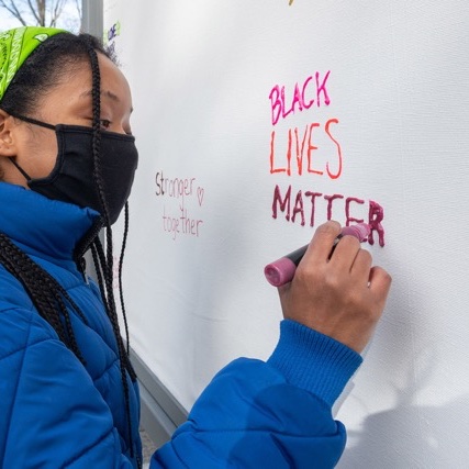A person wearing a mask and a blue jacket writes "black lives matter" on a whiteboard.
