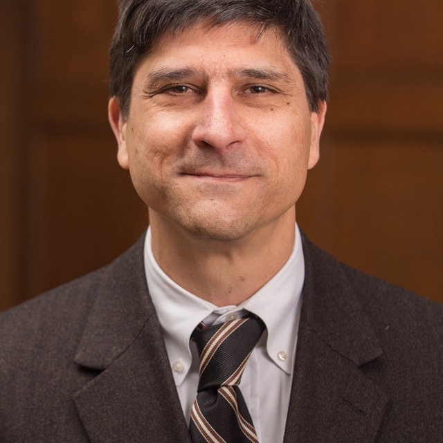 A person wearing a light colored shirt and brown jacket and tie smiles at the camera. They are in a room with wood paneling in the background.