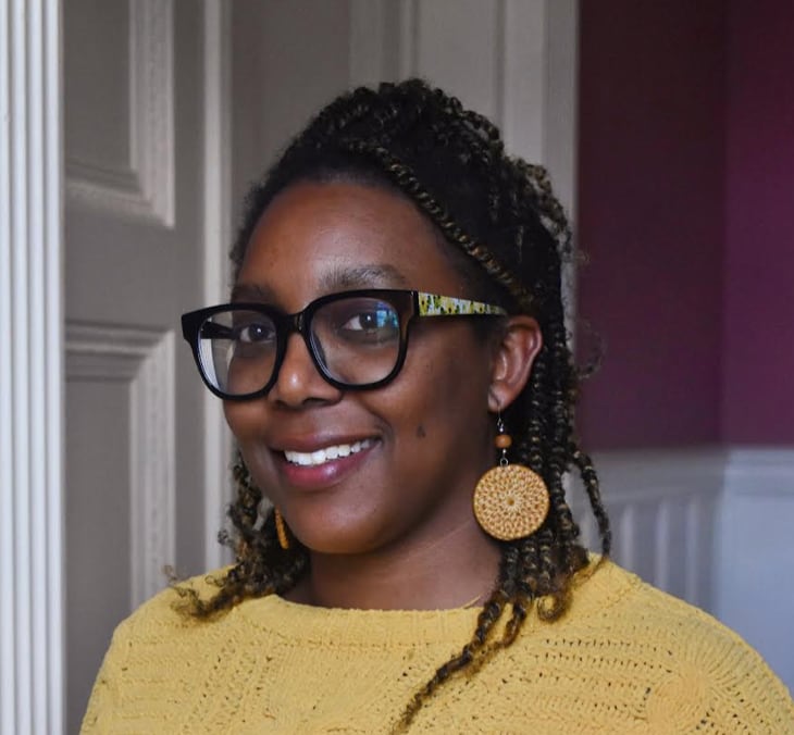 A person with long black braided hair, black-rimmed glasses, and a yellow shirt smiles at the camera.