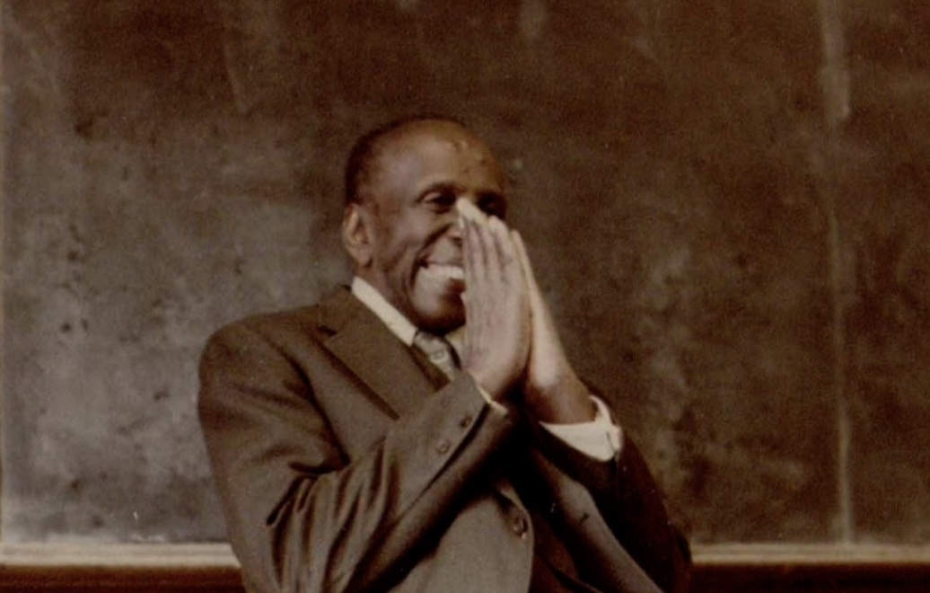 An old photograph of a person standing in front of a blackboard, smiling, hands pressed together in front of their face. The person is wearing a formal suit and tie.