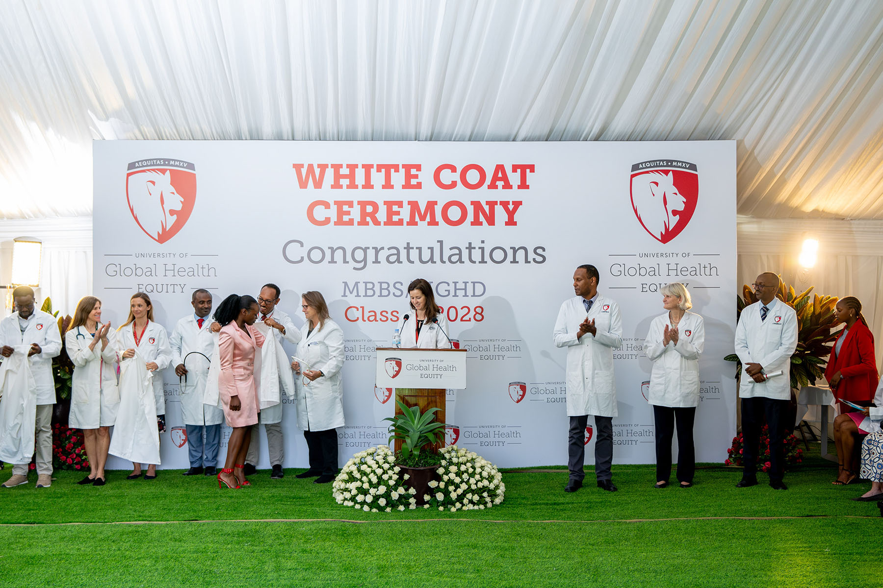 Person standing at a podium speaking flanked by people wearing lab coats. The tarp behind them reads, "White Coat Ceremony - Celebration."