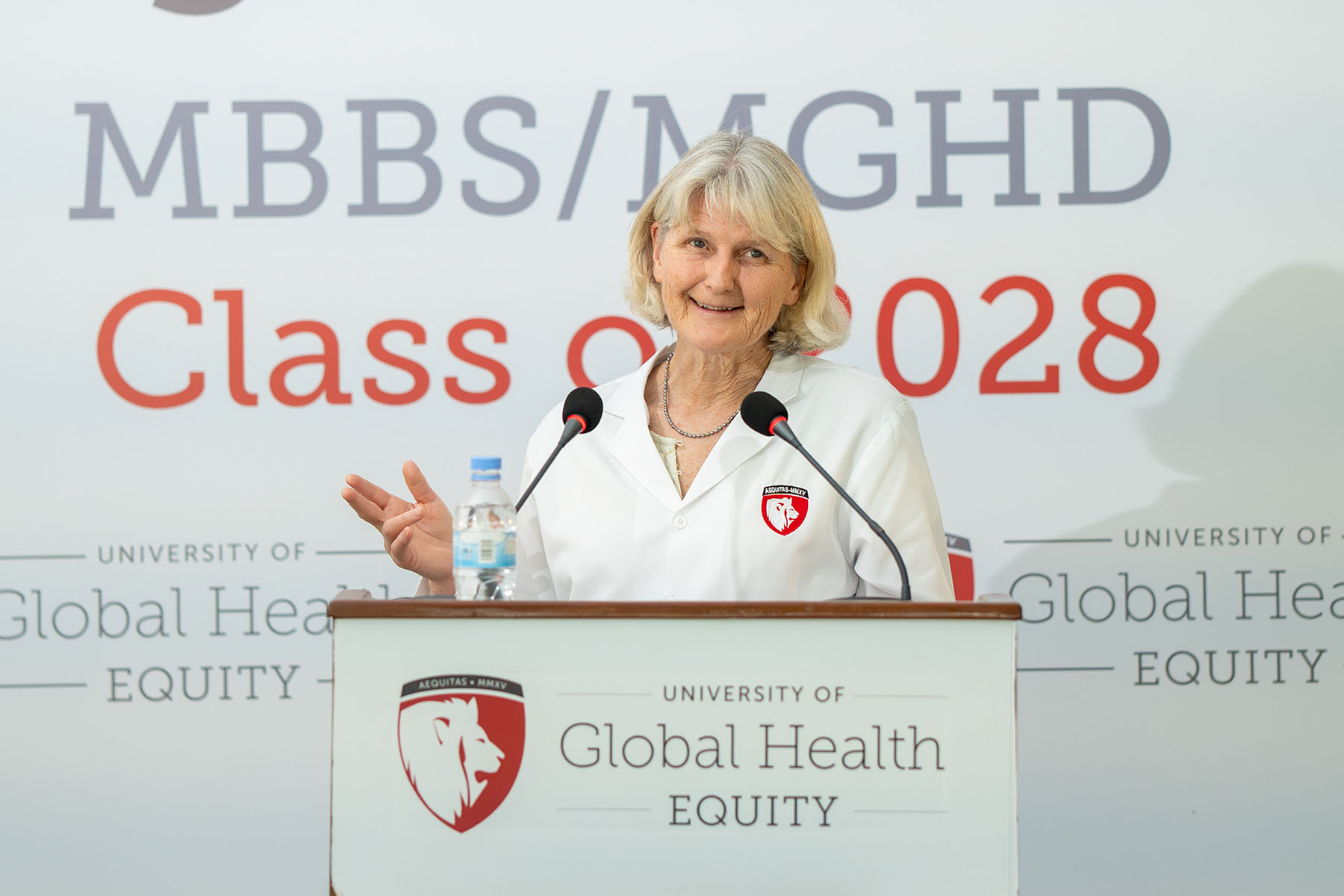 Pictured: Elizabeth Bradley. Person speaking at a podium with a white lab coat on. Behind the podium is a banner that reads, "MBBS/MGHD Class of 2028."