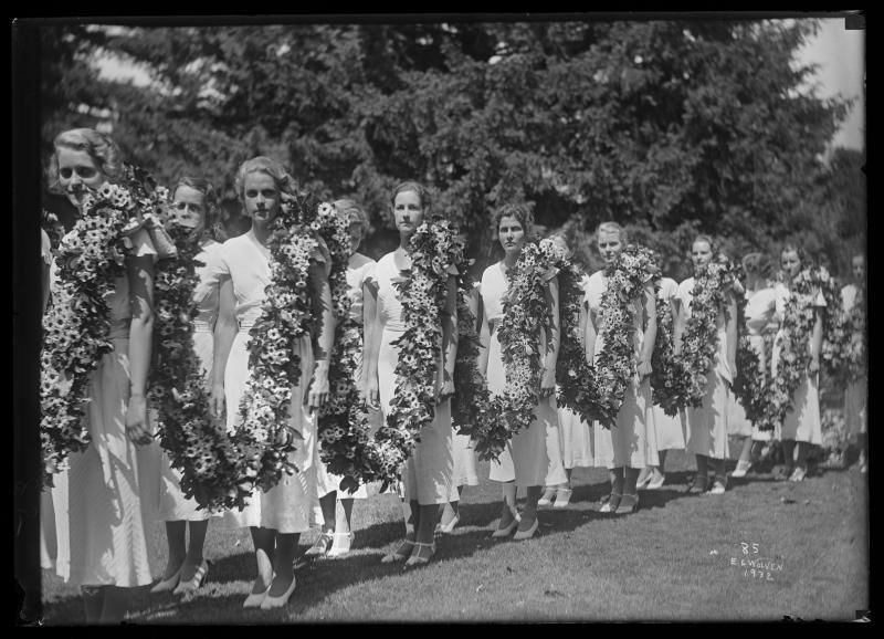 A black-and-white historical photo of a line of people wearing white dresses, standing outside on a lawn and holding a chain of flowers.