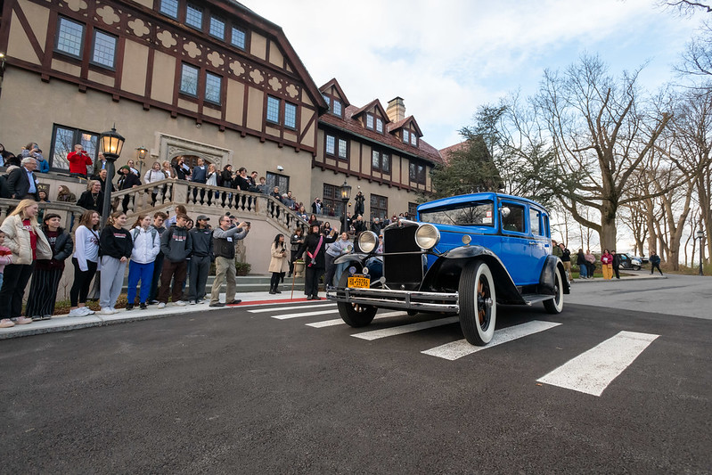 A classic blue vintage car is driven past a crowd of spectators gathered in front of a tudor-style building. The audience appears engaged and delighted by the vehicle.