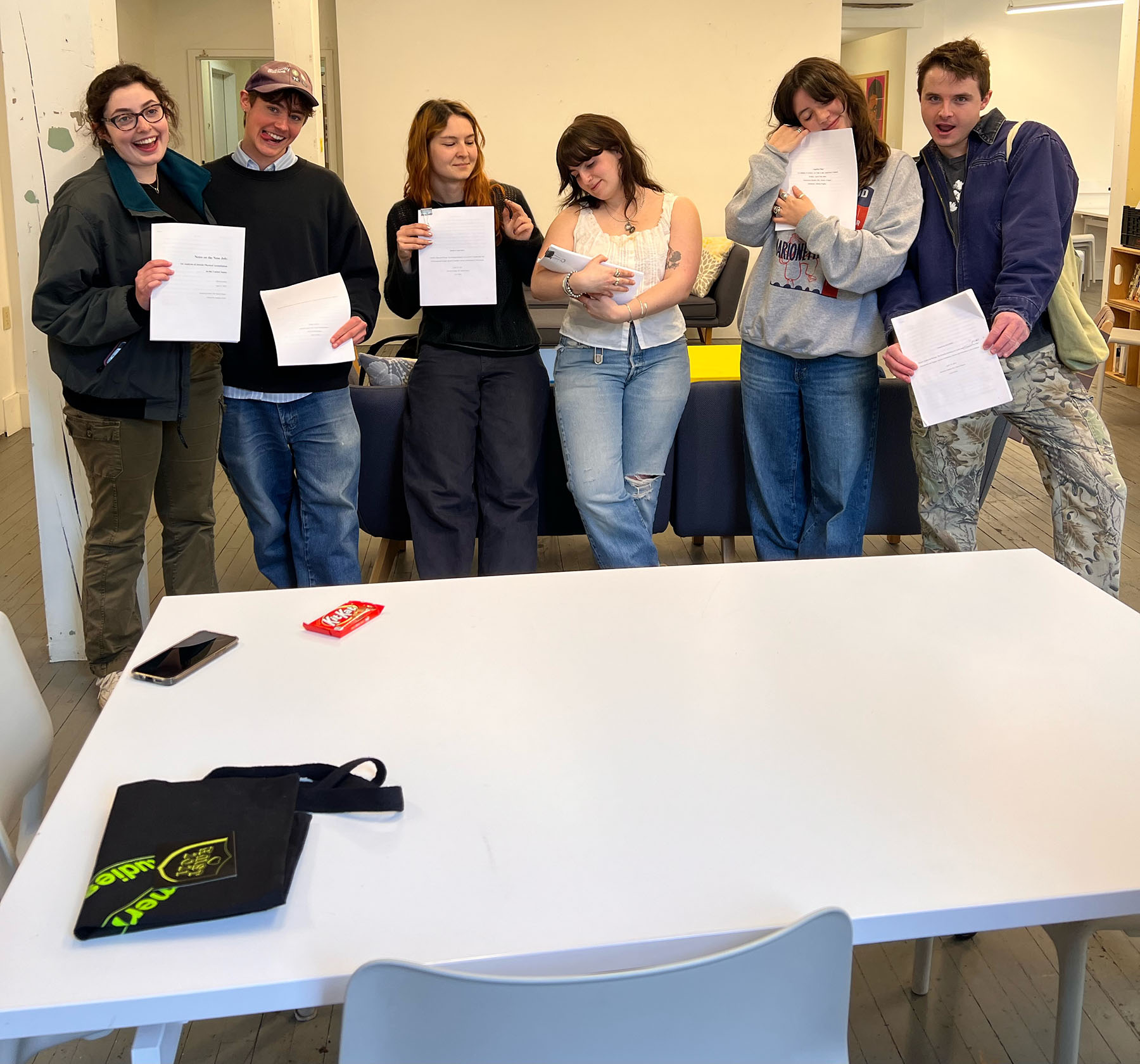 Six students holding pieces of paper standing in front of a table.