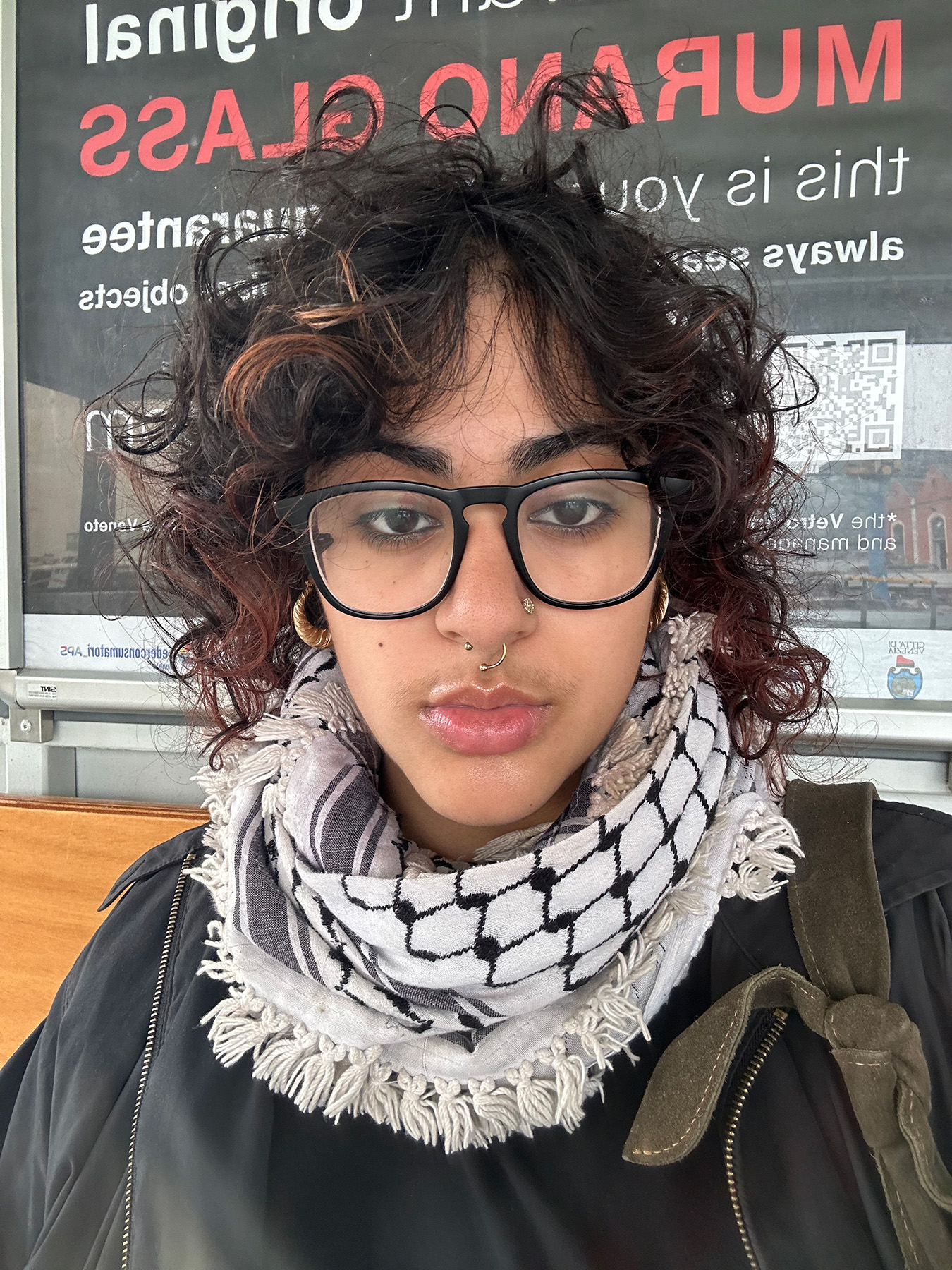 Person with black curly hair, dark glasses, scarf, and black jacket with both a nose ring and stud..