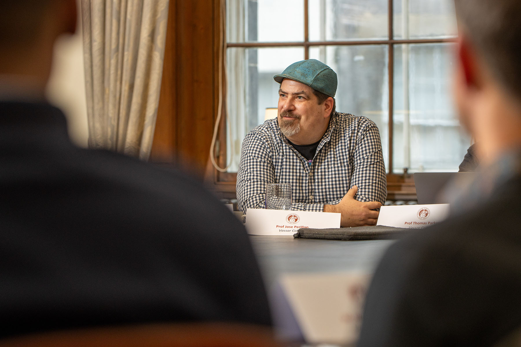 José Perillán sitting at a table, arms folded on top of it, wearing a green cap and black and white checkered patterned shirt.