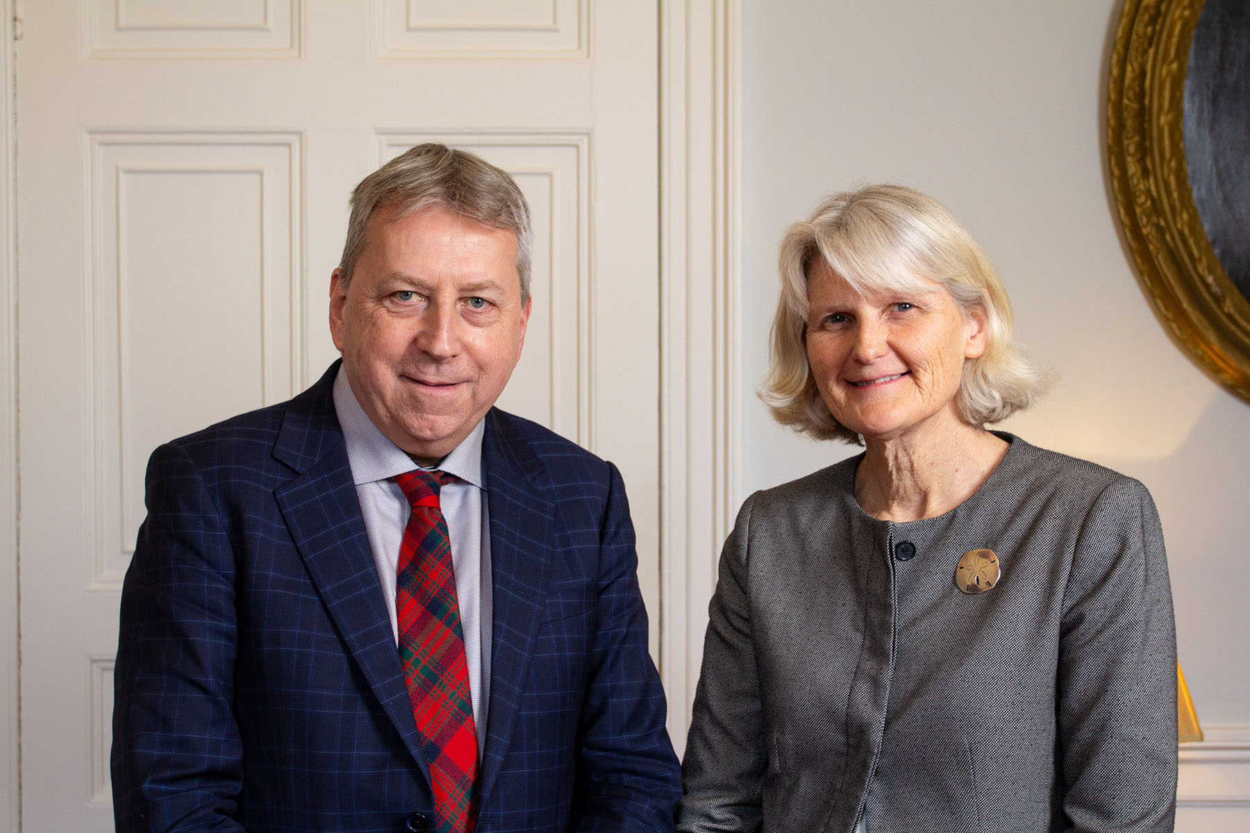 Sir Peter Mathieson, wearing a blue and white striped shirt, red plaid tie and navy blue jacket standing next to President Bradley who is wearing a grey shirt and sand dollar pin.