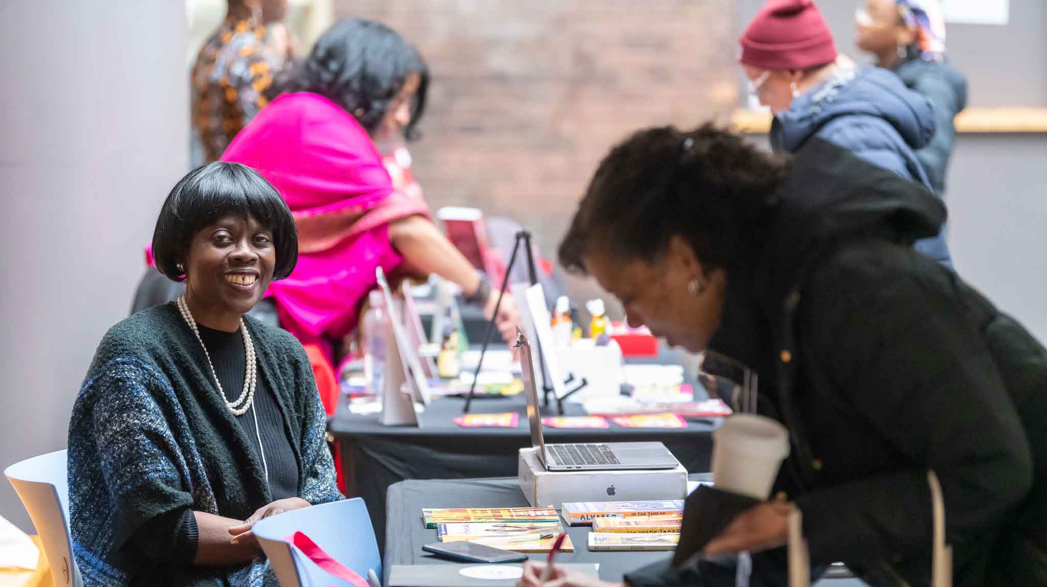 Person sitting at a table smiling at the camera while another person writes on a piece of paper on a table.