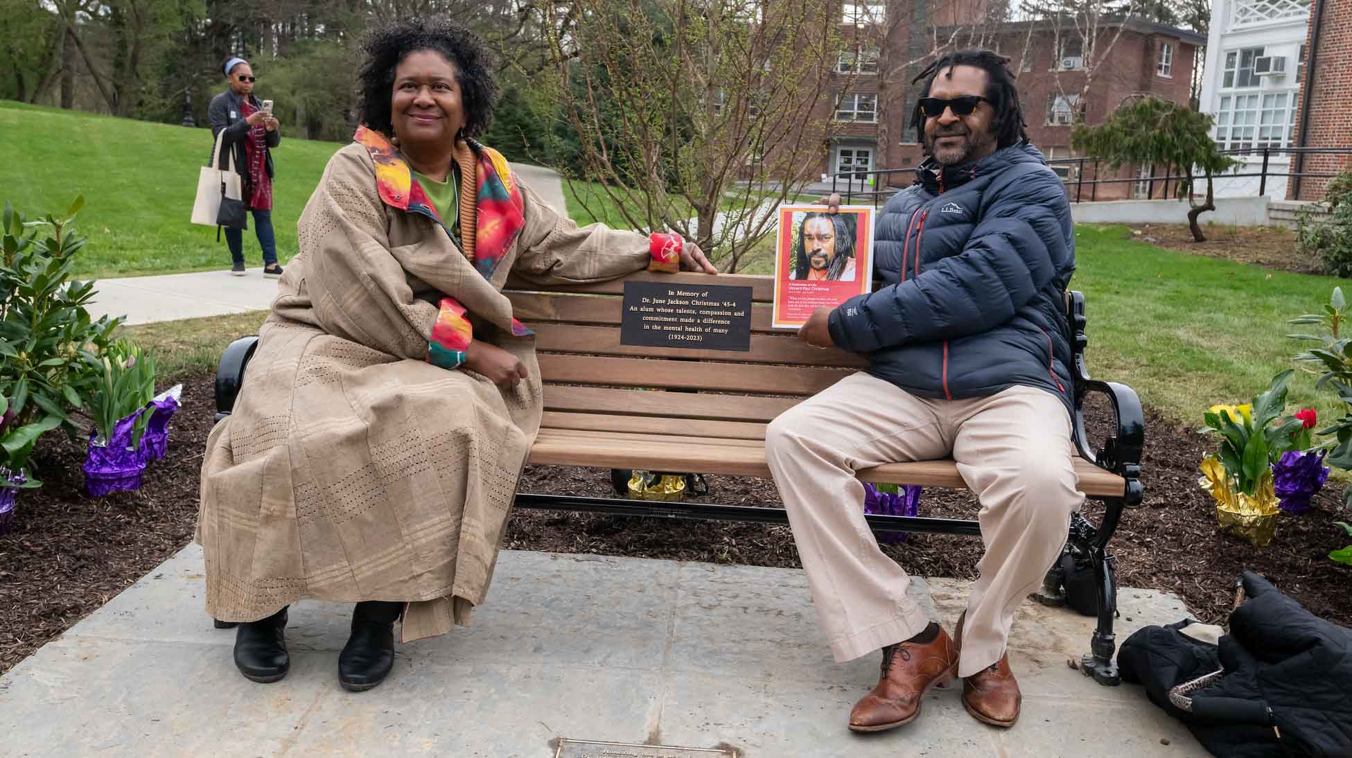 Two people seated on a bench, one person holding a portrait.