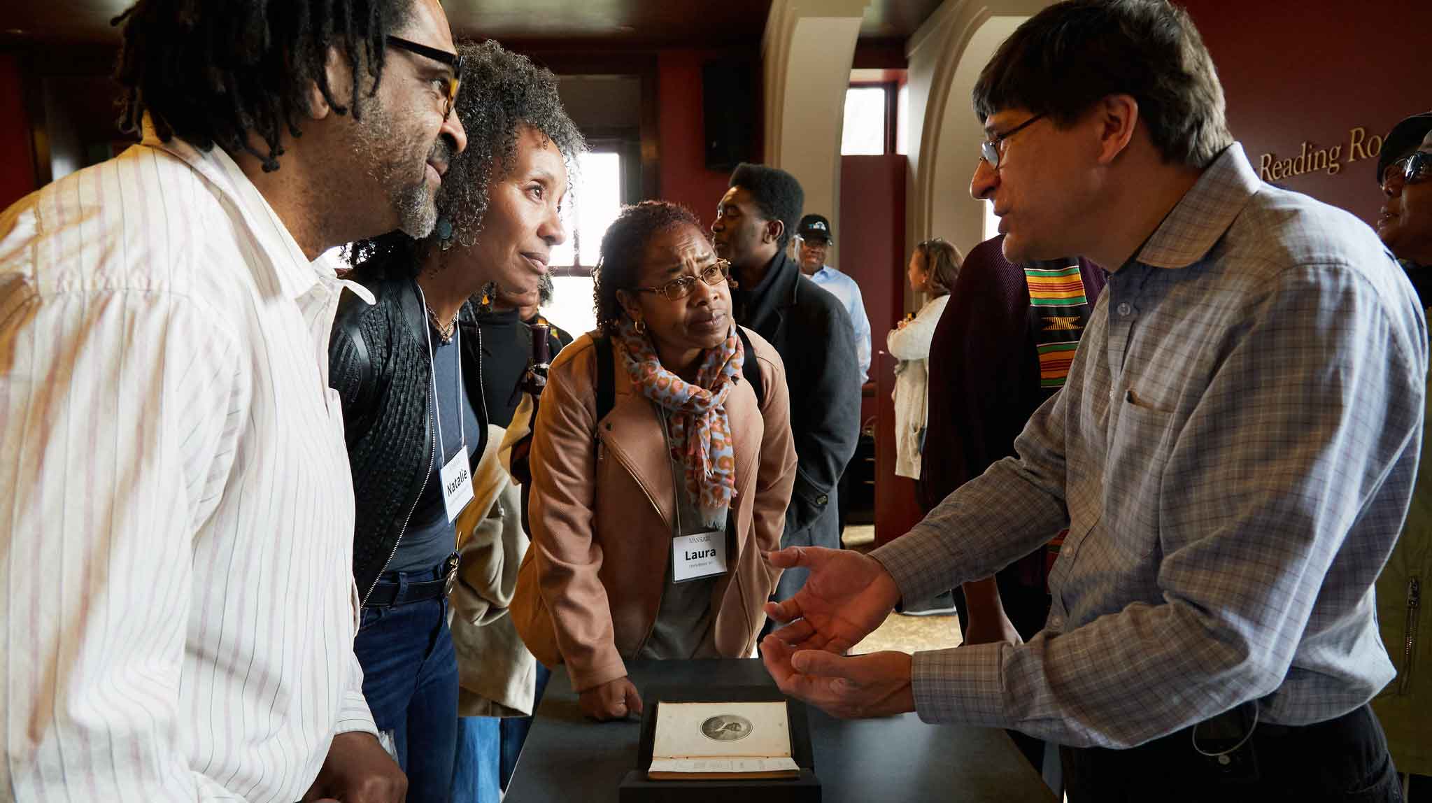 Group of people standing over a table with a book on it listening while one person talks.