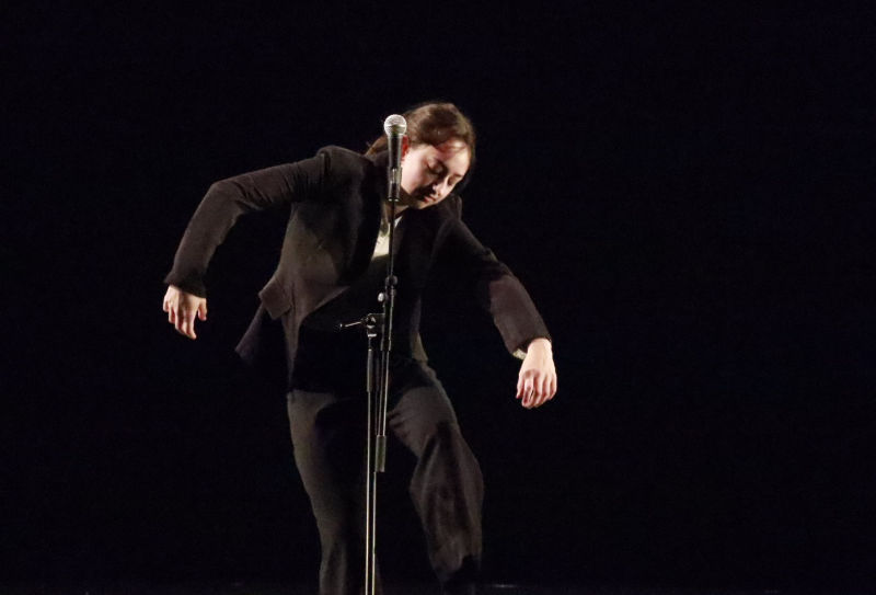 A person with long brown hair performs on a darkened stage in front of a microphone. The person is wearing all black clothing.