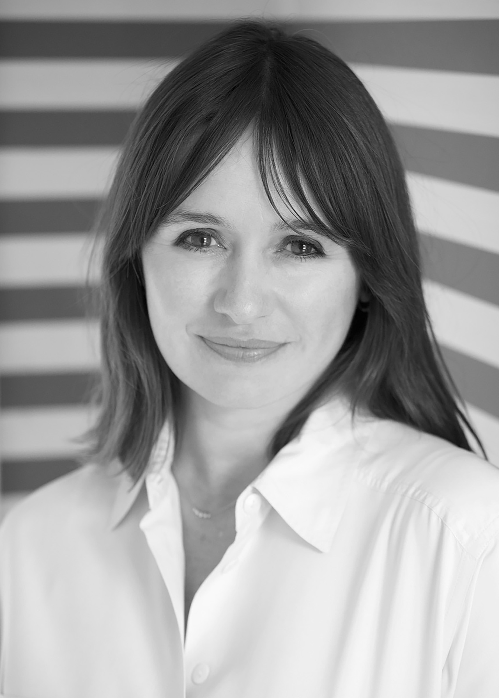 Black and white portrait of a person with shoulder-length hair, smiling slightly, wearing a white shirt, standing in front of a striped background.