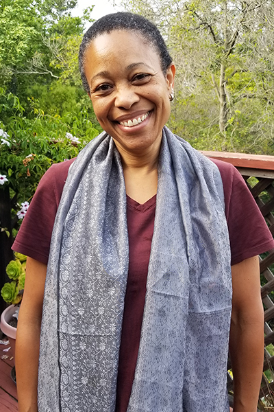 A portrait of Pam Harris standing outside on a deck with plants and trees all around.