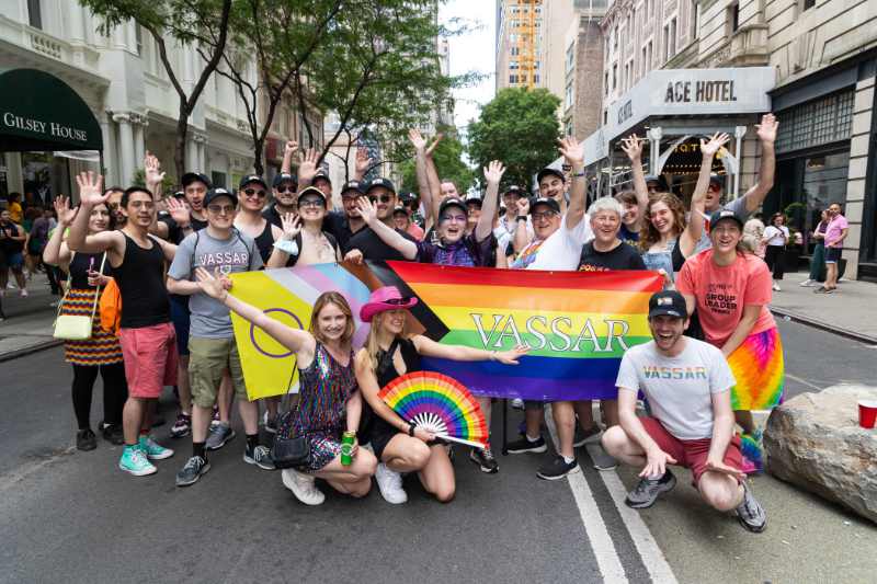 A group of smiling, waving people, some standing and some kneeling on the street in New York City, holding a rainbow banner with word "Vassar" on it.