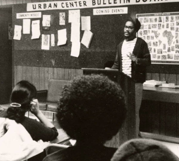 A black and white historical photo of a person standing at the front of a room, speaking to people. There is a bulletin board on the wall, with a banner that says "Urban Center Bulletin Board".