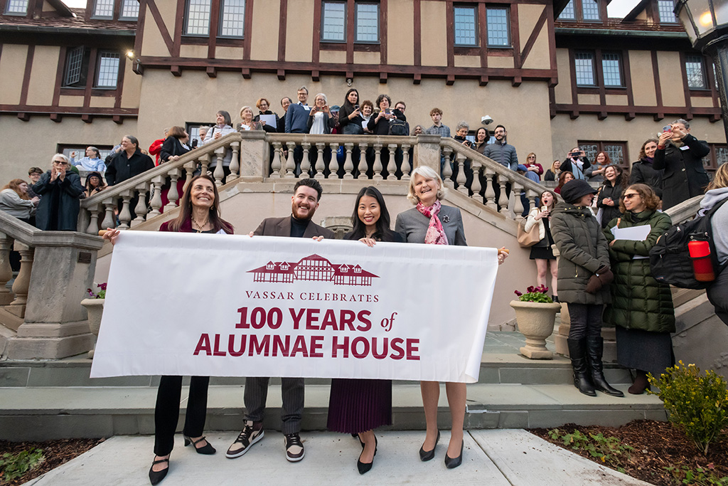 Four individuals holding a banner reading 'Vassar Celebrates 100 Years of Alumnae House' in front of the Tudor-style building with others standing behind them on stairs.