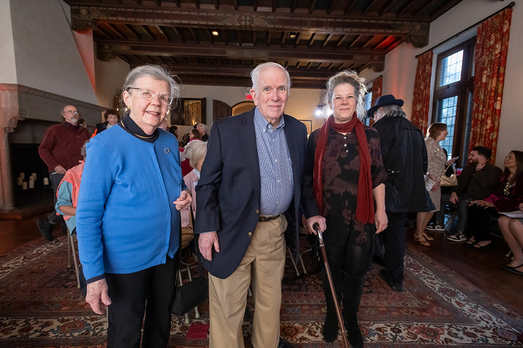 Three individuals stand together at an indoor event, smiling at the camera, with people milling in the background.