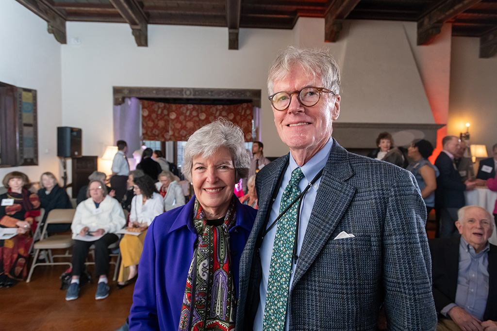 A couple stands together at an indoor event, smiling at the camera, with people milling in the background.