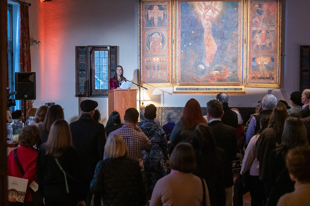 A woman speaks at a podium in front of an attentive audience in the historical living room.