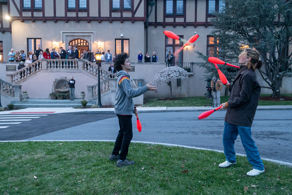 Two people juggling clubs outdoors, exchanging them mid-air, with an audience watching from the steps of Alumnae House during dusk.