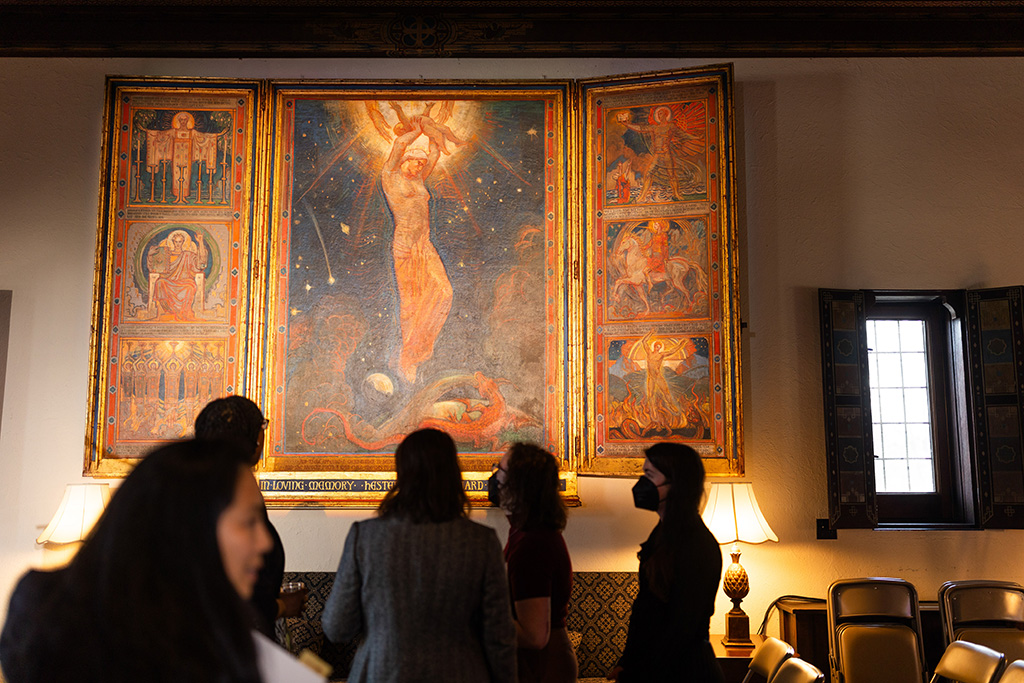 A group of individuals in the Alumnae House living room viewing a large art piece in a triptych format depicting a devotional image of a woman hovering against a star-filled sky in the center, flanked by smaller related narrative scenes from St. John's Book of Revelation.