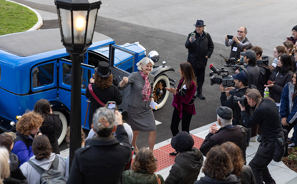 A tall woman wearing a grey suit with a red and white scarf stepping out of a classic blue car excitedly, greeted by a woman in a dark pink jacket, surrounded by a gathering of people including a camera operator capturing the scene.