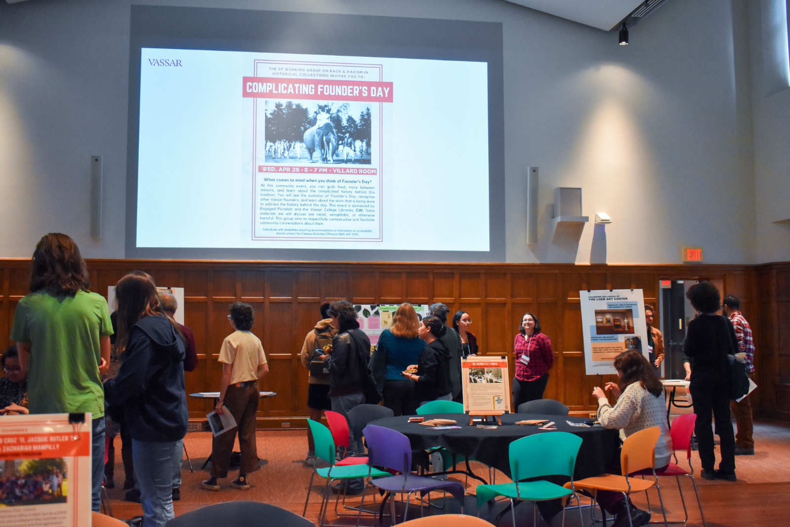 A group of people in a large, wood-paneled meeting room. Projected on a wall is a slide presentation. The slide shows a poster with the title “Complicating Founder’s Day”.