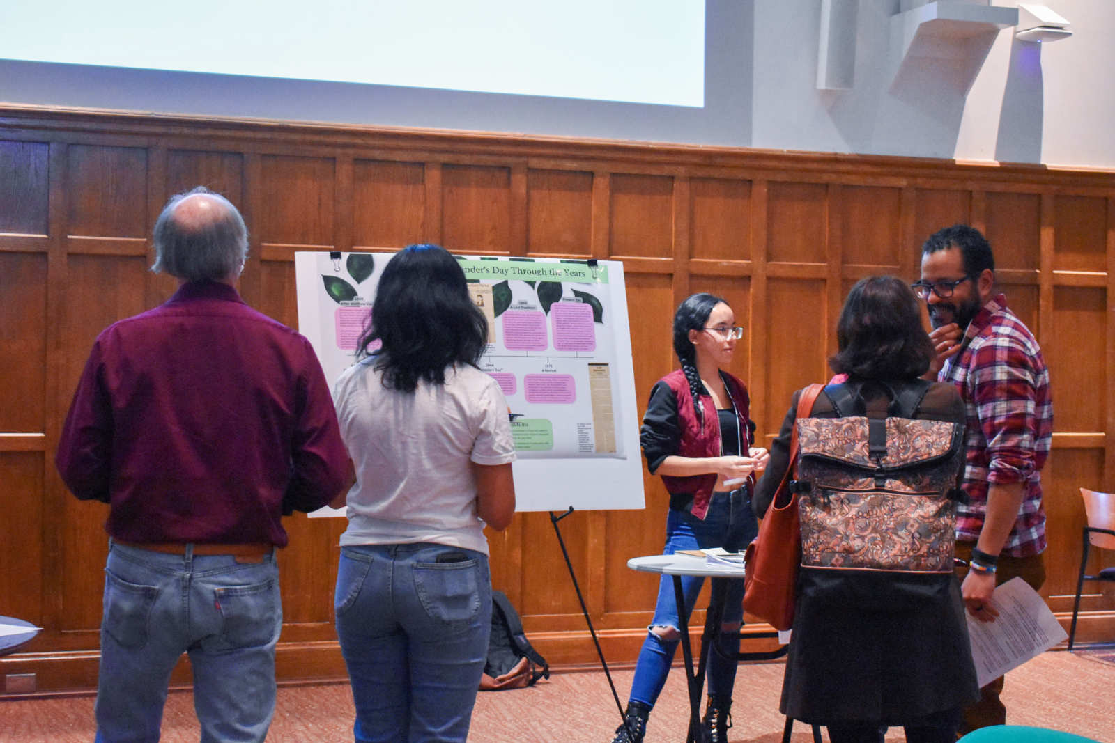 A group of people in a large, wood-paneled meeting room, looking at a large poster mounted on an easel.