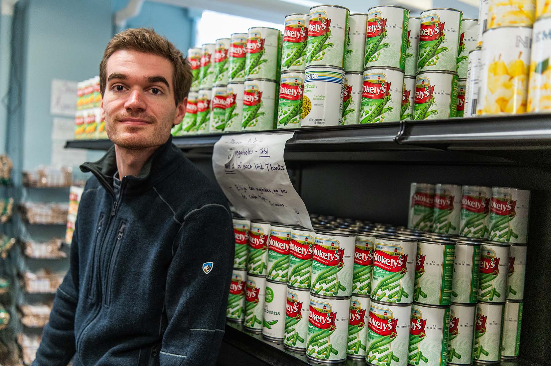 A person posing next to shelves filled with various canned food items.