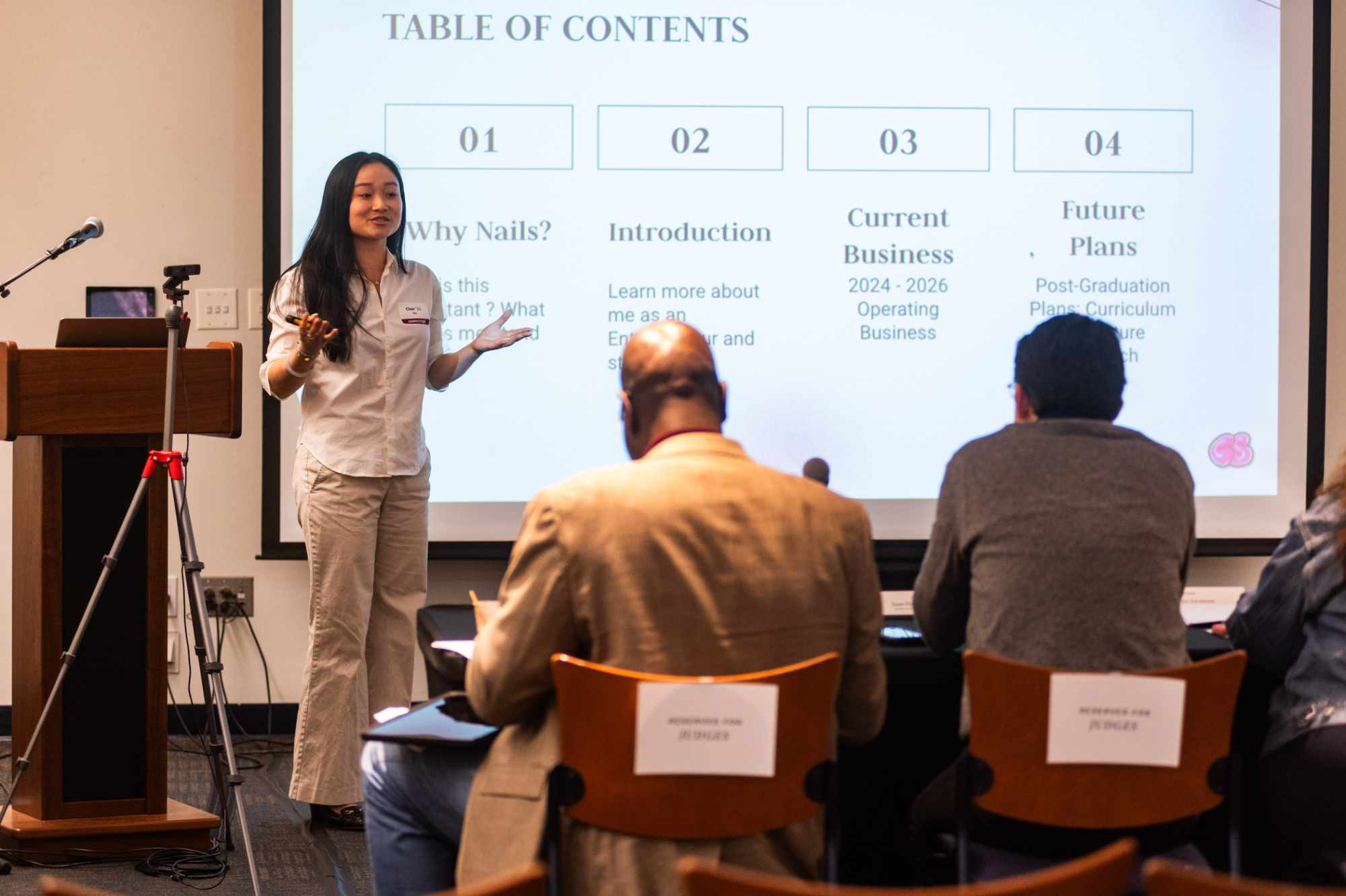 A person gives a presentation at the front of the room. Behind the person is a projected slide that reads “Table of Contents: Why Nails?; Introduction; Current Business; Future Plans”. Closest to the viewer are two people sitting in chairs, seen from the back.